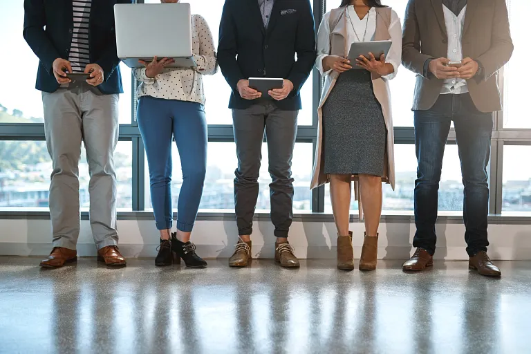 A group of five individuals standing indoors in front of large windows, shown from neck to feet. They are dressed in business casual attire and holding various devices, such as laptops, tablets, and smartphones. The polished floor reflects their footwear. The setting appears professional, possibly an office or conference space.